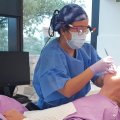 A person in a mask, safety glasses, gloves and hair net shines a light into the mouth of a prone man in pink clothing.